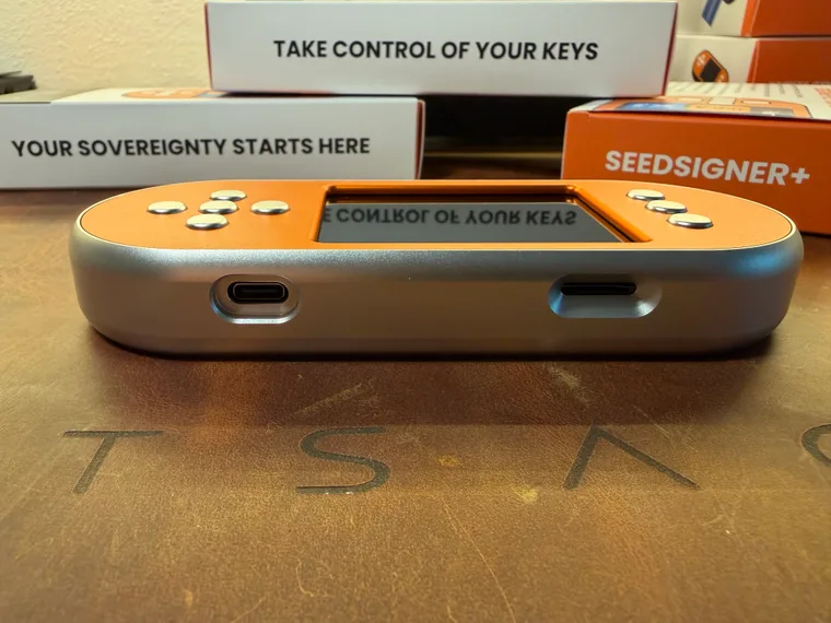 The bottom edge of the orange SeedSigner+ Premium, its orange front plate above a silver body, with the USB-C power port on the left and the microSD slot on the right