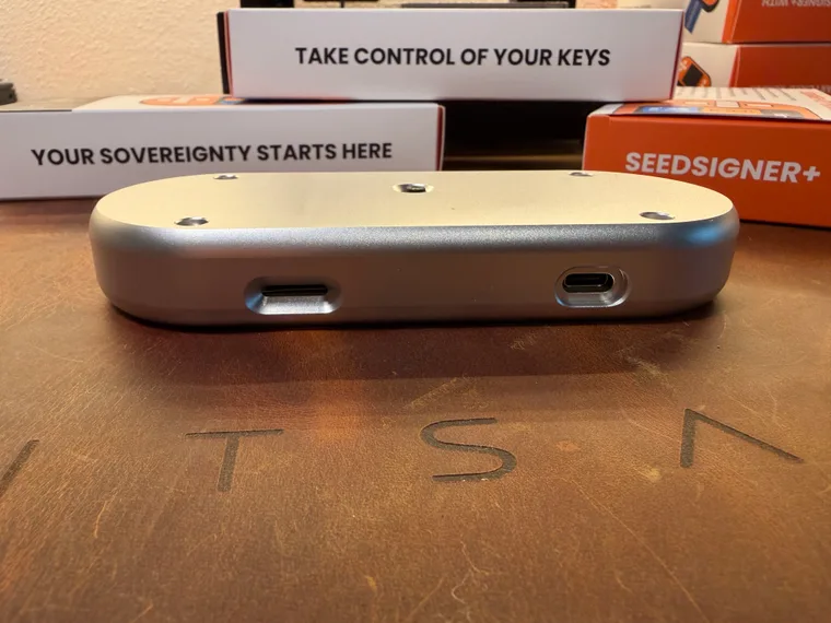 The orange SeedSigner+ Premium seen from below, where only its silver body shows, with the USB-C power port on the left and the microSD slot on the right, photographed in front of stacked SeedSigner+ retail boxes